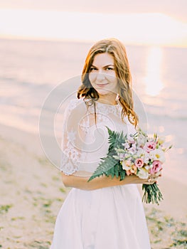 The lovely portrait of the smiling bride holding the wedding bouquet at the background of the sunset.