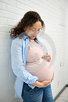 Lovely pregnant woman in 30 week of pregnancy, smiling and gentry stroking her belly, over white brick wall background.