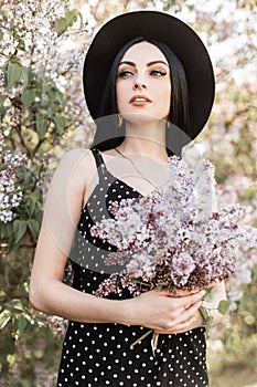 Lovely pretty woman in elegant hat in beautiful stylish dress stands with amazing color bouquet near lilac flowering trees in park