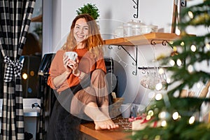 A lovely red-haired girl sits on the roofing cloth in a stylish cozy kitchen decorated for Christmas