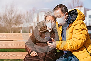 Lovely senior couple in medical masks using a smartphone while sitting on a bench in the park