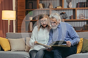 A lovely senior couple watching Memoirs photo album on sofa at home. Elderly family grandparents sitting together looking and