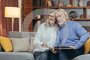 A lovely senior couple watching Memoirs photo album on sofa at home. Elderly family grandparents sitting together looking and