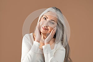 Lovely senior woman with beautiful long grey hair touching her face over brown studio background