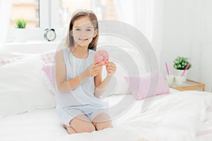 Lovely small girl holds delicious doughnut in hands, going to have breakfast, poses on bed in cozy white bedroom, looks at camera