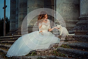 Lovely smiling bride with red hair is sitting on the old castle stairs and petting the pretty white cat.