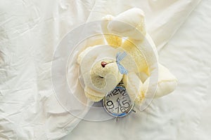 Lovely teddy bear on white bed background with alarm clock.