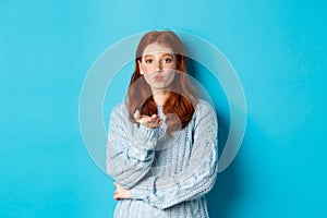 Lovely teen girl in sweater blowing air kiss, pucker lips and staring at camera, standing against blue background