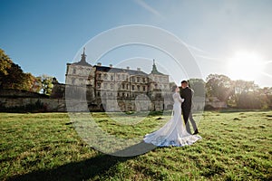 Lovely wedding couple walking in the sunlight near the old castle. Stylish handsome bride and groom in arms against the backdrop