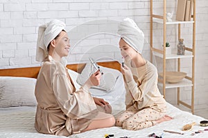 Lovely woman and her daughter in homewear and bath towels having fun with makeup, applying lipstick on bed at home