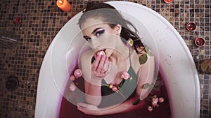 Lovely woman taking a bath with roses in spa. Pretty lady slowly puts rose in mouth looking up in camera. Cute girl