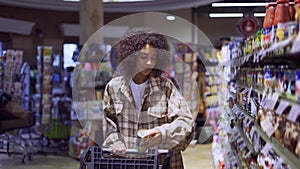 Lovely woman walking through aisle in supermarket with shopping cart looking at shelves