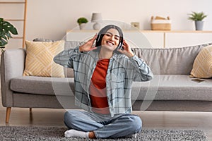 Lovely young Arab woman sitting on floor, wearing headphones, listening to music at home