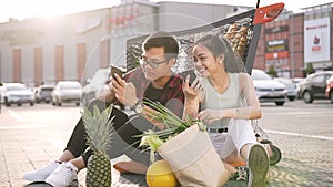 Lovely young asian man and woman in casual clothes sitting on the ground in front of supermarket and watching on their