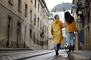 Lovely young couple with umbrella walking under rain on city street, back view