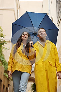 Lovely young couple with umbrella walking under rain on city street, low angle view