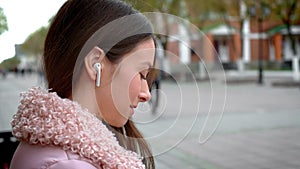 Lovely young girl is listening to music through wireless white headphones. Sits on a bench in the street in the fall