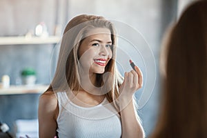 Sweet young girl looking in mirror and applying lipstick at home