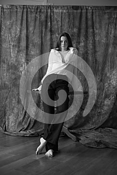 Lovely young girl model with dark long hair in a white shirt and black pants posing in the studio on a textile background.