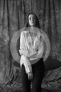 Lovely young girl model with dark long hair in a white shirt and black pants posing in the studio on a textile background.