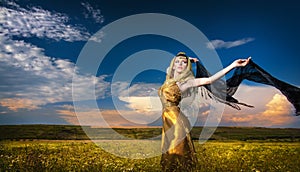 Lovely young lady posing dramatically with long black veil on green field. Blonde woman with cloudy sky in background - outdoor