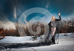 Lovely young lady posing dramatically with long black veil in winter scenery. Blonde woman with cloudy sky in background - outdoor