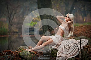 Lovely young lady sitting near river in enchanted woods. Sensual blonde with white clothes posing provocatively in autumnal park.