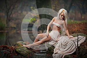 Lovely young lady sitting near river in enchanted woods. Sensual blonde with white clothes posing provocatively in autumnal park.