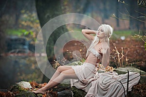 Lovely young lady sitting near river in enchanted woods. Sensual blonde with white clothes posing provocatively in autumnal park.