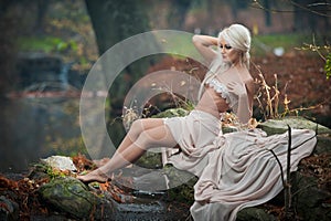 Lovely young lady sitting near river in enchanted woods. Sensual blonde with white clothes posing provocatively in autumnal park.