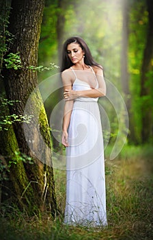 Lovely young lady wearing an elegant long white dress enjoying the beams of celestial light on her face in enchanted woods. Long