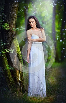 Lovely young lady wearing an elegant long white dress enjoying the beams of celestial light on her face in enchanted woods. Long