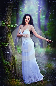 Lovely young lady wearing an elegant long white dress enjoying the beams of celestial light on her face in enchanted woods. Long