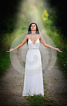 Lovely young lady wearing an elegant long white dress enjoying the beams of celestial light on her face in enchanted woods. Long