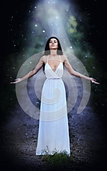 Lovely young lady wearing an elegant long white dress enjoying the beams of celestial light on her face in enchanted woods. Long