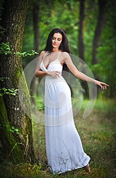 Lovely young lady wearing an elegant long white dress enjoying the beams of celestial light on her face in enchanted woods. Long