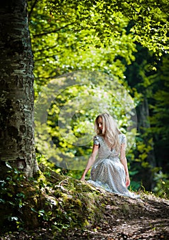 Lovely young lady wearing elegant white dress enjoying the beams of celestial light on her face in enchanted woods. Pretty blonde