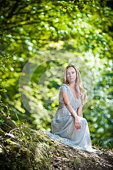 Lovely young lady wearing elegant white dress enjoying the beams of celestial light on her face in enchanted woods. Pretty blonde