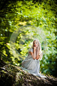 Lovely young lady wearing elegant white dress enjoying the beams of celestial light on her face in enchanted woods. Pretty blonde