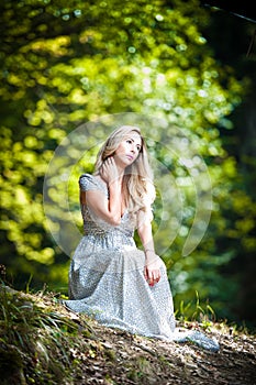 Lovely young lady wearing elegant white dress enjoying the beams of celestial light on her face in enchanted woods. Pretty blonde