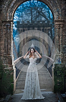 Lovely young lady wearing elegant white dress and silver tiara posing on ancient bridge, ice princess concept. Pretty brunette