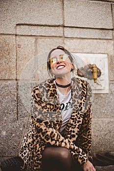 Lovely young laughing brunette woman posing outdoor on old stone fountain background. Wearing stylish yellow glasses, trendy