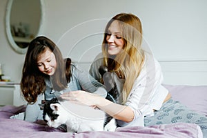Lovely young mother and cute school kid girl cuddling together with cat in bed in morning. Happy family of young single