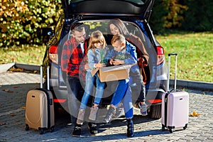 Lovely young parents with their cute kids sitting in the trunk and holding carton box with plants and other home things