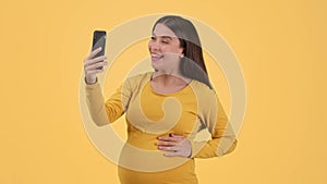 Lovely young pregnant woman video chatting with friends via smartphone, waving hand and sending blow kiss to device