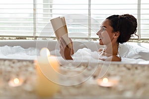 Lovely young woman lying in foamy bath, reading book in relaxing atmosphere with candles, indoors. Copy space