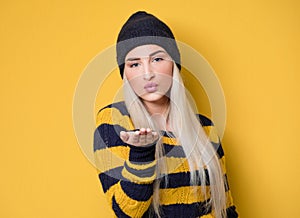 Lovely young woman send kiss to you, model wearing woolen cap and sweater,  on yellow background. Beautiful lovely woman