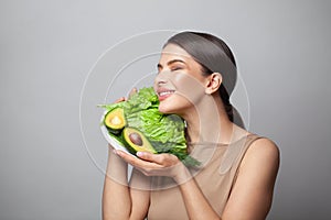 Lovely young woman smiling cheerfully and showing avocado and green lettuce on plate.