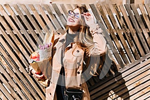 Lovely young woman in stylish beige coat enjoys sunlight closing eyes after shopping. Pretty girl standing outside face