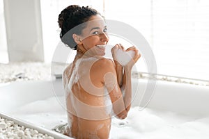 Lovely young woman taking hot bubble bath, making foamy heart with her hands, enjoying spa procedure at home